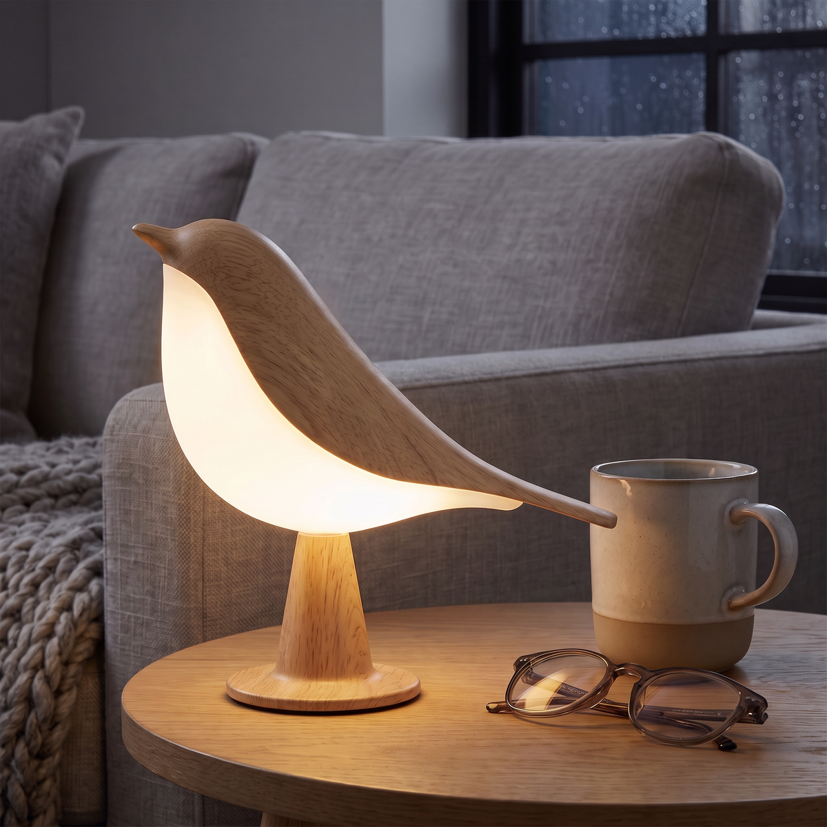 Lampe Oiseau bois clair sur une table d'appoint près du canapé, avec un mug et des lunettes